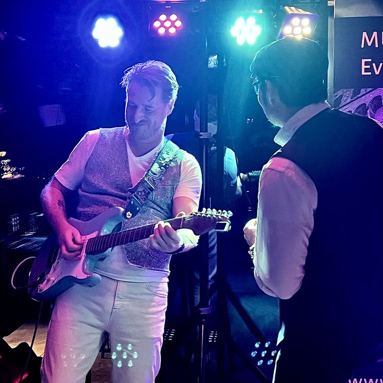 Gitarrist spielt E-Gitarre im blauen Scheinwerferlicht, daneben ein italienischer Sänger